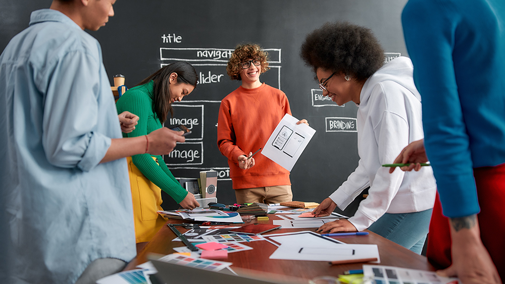 Young cheerful designer is showing some sketches to his colleagues while having a meeting in the modern office. Working together. Teamwork. Web design concept